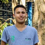 Young man smiling outdoors wearing a grey V-neck T-shirt with logos from Raleigh Costa Rica and Youth for Green Growth.